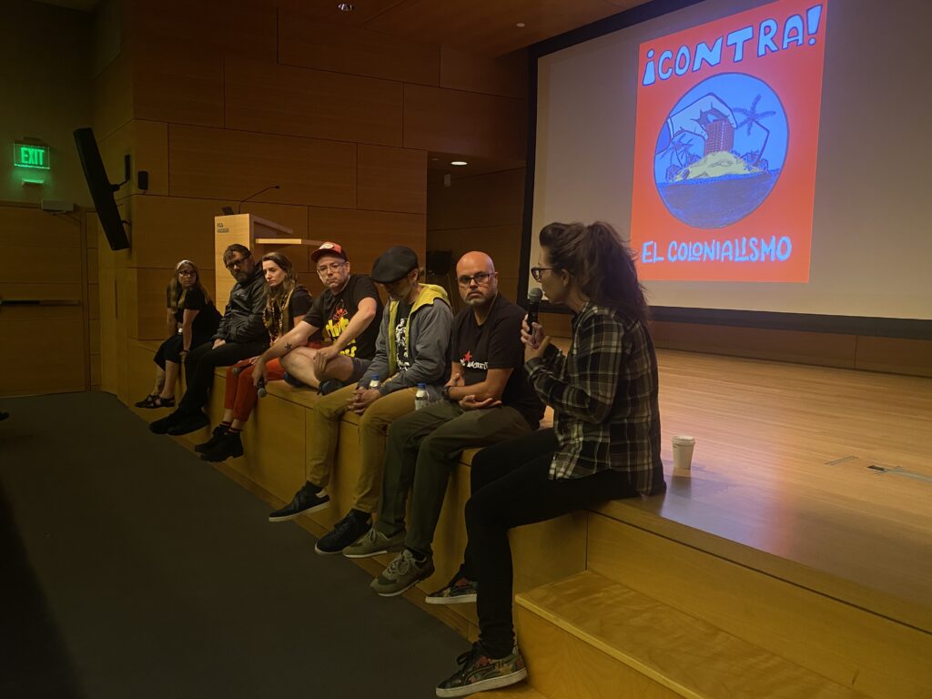 A group of light skinned people sit on a stage. One person, a femme with light hair in a pony tail, is speaking into a microphone. In the background there is a screen with a red poster that reads "!Contra! El Colonialismo